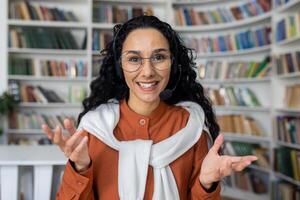 webbkamera se uppkopplad ringa upp, ung kvinna studerande talande till vänner avlägset, latinamerikan kvinna inuti akademisk bibliotek på skrivbord studerar och framställning för tentor. foto