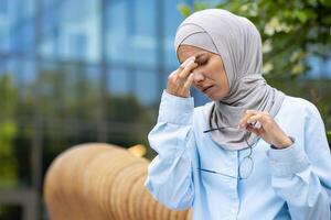 en medelålders affärskvinna i en ljus blå hijab och skjorta, innehav henne glasögon och rörande henne tempel, utseende betonade eller sjuk mot ett urban bakgrund. foto