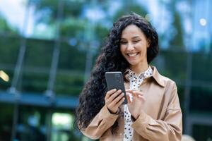 ung skön latinamerikan kvinna gående i de stad, företag kvinna innehav telefon i händer använder sig av smartphone app, kvinna leende belåtet och Lycklig utanför kontor byggnad med lockigt hår foto