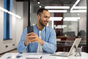 leende ung arab man arbetssätt i kontor på bärbar dator, Sammanträde på tabell och ser på skärm, innehav och använder sig av mobil telefon. foto