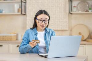 uppkopplad handla. en ung skön asiatisk kvinna i glasögon sitter på Hem på de tabell med en vit bärbar dator, innehar en kreditera kort i henne händer. köper och order varor på de internet. foto