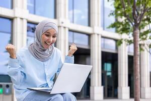 en glad muslim kvinna i en hijab höjer henne nävar i Framgång medan använder sig av en bärbar dator utomhus nära modern kontor byggnader. foto