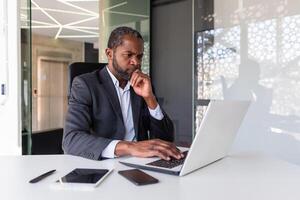 tänkande mogna vuxen afrikansk amerikan affärsman på arbetsplats, finansiär chef investerare med bärbar dator tänkande handla om finansiell lösningar ny projekt inuti kontor. foto