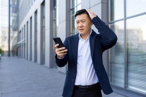 en ung asiatisk man i en företag kostym är stående utanför ett kontor Centrum, ser chockade och orolig på de telefon skärm, och innehav hans huvud. foto