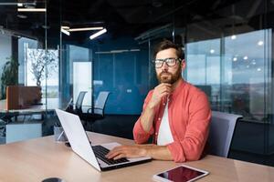 allvarlig och fokuserade mogna affärsman i röd skjorta bär glasögon ser på kamera, ung man på arbete skriver på bärbar dator tangentbord, programmerare utvecklare porträtt på arbete. foto