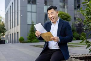 ett extatisk affärsman i en kostym sitter utanför ett kontor byggnad, glatt läsning en brev med en ljus leende. foto
