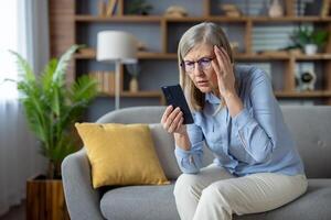 ett äldre kvinna sitter på en soffa, uttrycker förvirring medan ser på henne smartphone, eventuellt som handlar om med teknologi frågor. foto