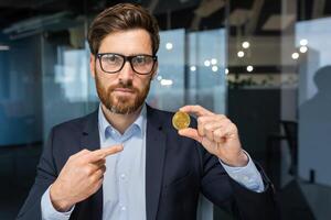 allvarlig och fokuserade affärsman ser på kamera, mogna framgångsrik investerare innehav bitcoin mynt kryptovaluta och pekande, skäggig man arbetssätt inuti modern kontor. foto