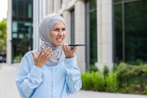 irriterad kvinna i hijab talande med telefon högtalare och grimaserande eftersom av ljud på stadens centrum gata. betonade vuxen kvinna kämpar med använder sig av mobil digital grej och förlorar förbindelse. foto