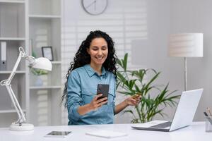 glad latina affärskvinna använder sig av smartphone och innehav kreditera kort, arbetssätt från en välorganiserad Hem kontor med bärbar dator och grön växter runt om. foto