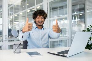 en ung man i en ljus blå skjorta Sammanträde på en skrivbord i en ljus kontor miljö, entusiastiskt ger två tummen upp medan leende, porträtterar Framgång och tillfredsställelse på arbete. foto