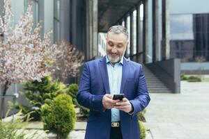 en senior gråhårig man i en företag kostym är stående nära ett kontor Centrum med en telefon i hans händer. gläds, firar vinnande de lotteri, spelar på de telefon. foto