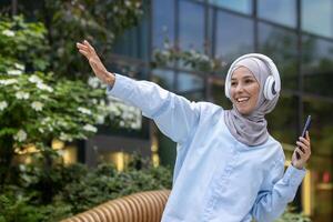 ung muslim kvinna studerande utanför universitet campus, Lycklig dans utanför byggnad, kvinna i hijab lyssnande till musik använder sig av telefon och hörlurar, Lycklig leende. foto