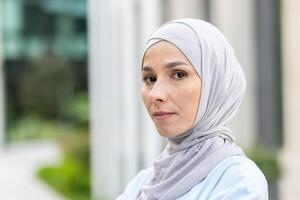 porträtt av en ung muslim kvinna bär en hijab, ser självsäkert på de kamera med ett kontor byggnad bakgrund, förmedla professionalism och elegans. foto