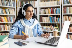en kvinna studerande bär hörlurar koncentrat på henne bärbar dator medan studerar i en bibliotek. hon är omgiven förbi böcker och har kaffe närliggande, indikerar en lång studie session. foto
