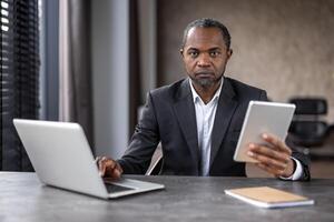fokuserade afrikansk amerikan affärsman använder sig av en bärbar dator och läsplatta samtidigt i en modern kontor miljö. han visas allvarlig och tillägnad till hans arbete. foto