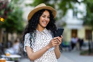 en skön ung kvinna promenader genom de kväll stad i en hatt, en leende latin amerikan kvinna innehar en smartphone i henne händer. en turist med lockigt hår typer en meddelande och bläddrar uppkopplad sidor på de telefon. foto