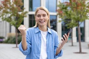 en glad ögonblick fångad som en person firar Framgång eller Bra Nyheter utomhus, innehav upp en näve i en triumferande gest med en smartphone i de Övrig hand, skildrar seger, prestation, och lycka. foto