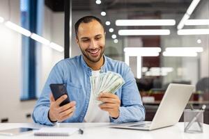 Lycklig affärsman i en tillfällig blå skjorta innehav kontanter och en mobil telefon, känsla framgångsrik på arbete i en modern kontor. foto
