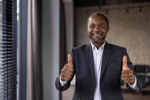 glad afrikansk amerikan affärsman i en kostym ger en dubbel- tummen upp, symboliserar godkännande och Framgång i ett kontor miljö. foto