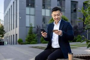 leende ung asiatisk man Sammanträde på bänk i företag kostym, använder sig av mobil telefon och kreditera kort. foto
