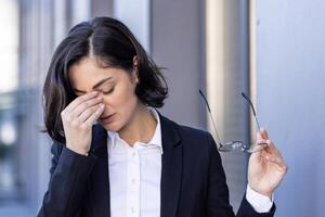 ung kvinna trött med huvudvärk, affärskvinna utanför kontor byggnad gnuggar ögon, yr, överarbetad arbetstagare utomhus i företag kostym stänga upp. foto