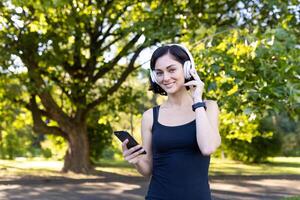 leende kvinna med smartphone och hörlurar njuter en musik ha sönder i natur under en kondition rutin- på en solig dag. foto