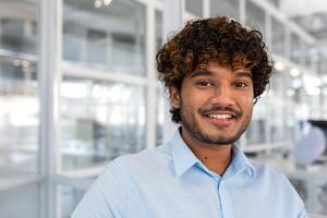 ung leende indisk programmerare stänga upp leende och ser på kamera, porträtt av man med lockigt hår och blå skjorta inuti kontor på arbete, affärsman entreprenör med skägg arbetssätt på projekt. foto