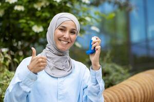 en muslim kvinna i en hijab innehar ett astma inhalator i en parkera, ger en tummen upp för hälsa och friskvård. foto