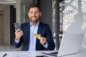 porträtt av en Lycklig och leende ung affärsman, bankman Sammanträde i en kostym i de kontor på de tabell med en bärbar dator, innehav en telefon och en kreditera kort, ser på de kamera. foto