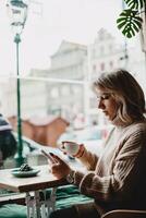 kvinna njuter kaffe och telefon på Kafé tabell. en kvinna sitter på en Kafé tabell, innehav en kopp av kaffe och kontroll henne telefon. en inlagd växt lägger till charm till de mysigt Kafé atmosfär. foto