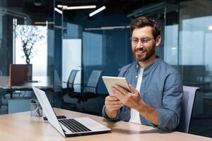 framgångsrik affärsman i en skjorta användningar en läsplatta dator, en man sitter på en skrivbord leende och Lycklig, en programmerare tester en ny Ansökan. foto