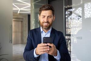 närbild Foto av en ung affärsman leende och nervöst använder sig av en mobil telefon medan Sammanträde inuti de kontor.