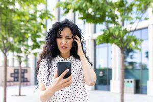 angelägen ung kvinna med lockigt hår ser på telefon skärm med en bekymrad uttryck, stående utomhus nära kontor byggnader. foto