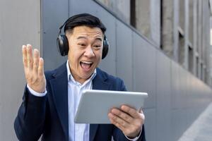 Lycklig ung asiatisk manlig affärsman stående i hörlurar utanför kontor Centrum, ser på läsplatta skärm och Lycklig med Nyheter och Framgång. foto