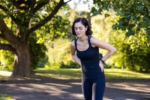 aktiva och sportig ung kvinna stående i de parkera böjd över, innehav henne händer Bakom henne tillbaka, känsla svår smärta och Trötthet efter joggning och Träning. foto