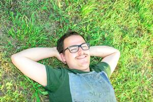 ung man med glasögon lied på gräs njuter semester efter studerar mycket Lycklig foto