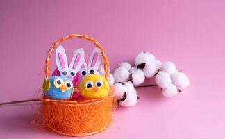 en gren av vit bomull, leksak ägg med fåglar i ett orange korg och påsk kaniner på en rosa bakgrund. foto