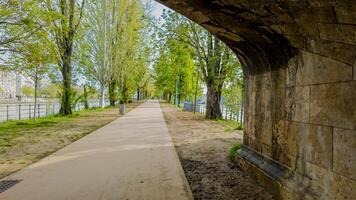 lugn flodstrand promenad med lång grön träd i vår, under ett gammal sten bro båge, idealisk för begrepp relaterad till fritid och urban natur foto