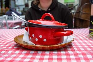 traditionell kål soppa i de röd kasta järn pott. de skål är eras på de keramisk tallrik med vit vävnad. retro stil pläd tabell trasa. utomhus restaurang. människor i de suddig bakgrund. foto