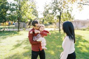 pappa kyssar en liten flicka i hans vapen Nästa till en stående mamma i de parkera foto