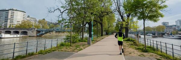 man joggning förbi de flod i ett urban parkera, med tidigt vår lövverk och stad byggnader i de bakgrund relaterad till kondition och utomhus- aktiviteter foto