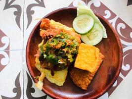 indonesiska mat. kyckling penyet med chili sås med tofu, tempeh, gurka, ägg och Övrig färsk grönsaker på trä tallrik foto