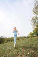 ett atletisk flicka utför en jogga i de parkera. skön blond caucasian kvinna i blå tajt träningsoverall. blond flicka på ett utomhus- Träning session foto