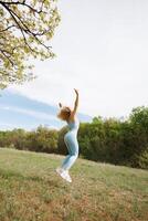 ett atletisk flicka utför de övning av Hoppar upp med henne vapen Uppfostrad. skön blond caucasian kvinna i blå tajt träningsoverall. blond flicka på ett utomhus- Träning session foto