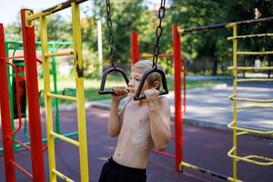en pojke med ett atletisk bygga drar han själv upp på hängande skenor. gata träna på en horisontell bar i de skola parkera. foto