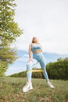 en flicka övningar med en sudd band på henne på henne ben. skön blond caucasian kvinna i blå tajt träningsoverall. blond flicka på ett utomhus- Träning session foto