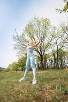 en flicka idrottare utför vikt förlust övningar i de parkera med en sudd band. skön blond caucasian kvinna i blå tajt träningsoverall. blond flicka på ett utomhus- Träning session foto