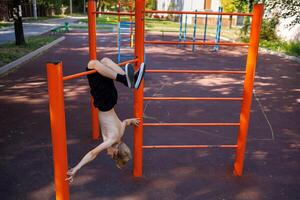 en tonåring av atletisk bygga hänger upside ner med hans fötter på de bar. gata träna på en horisontell bar i de skola parkera. foto