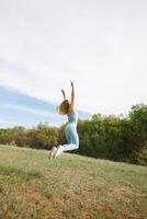 ett atletisk flicka utför de övning av Hoppar upp med henne vapen Uppfostrad. skön blond caucasian kvinna i blå tajt träningsoverall. blond flicka på ett utomhus- Träning session foto